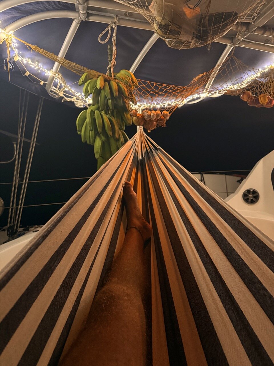 A person relaxing in a striped hammock on the deck of the luxury catamaran SV Monika, with hanging bananas and string lights overhead, enjoying a peaceful evening at sea.