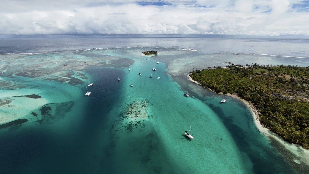 Aerial view of SV Monika, a luxury catamaran, sailing through clear blue waters surrounded by tropical islands and coral reefs, showcasing an idyllic Caribbean paradise.