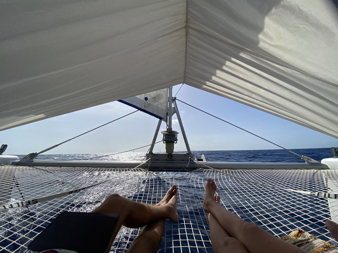 A view from inside the SV Monika luxury catamaran, showing the deck with two people relaxing and enjoying the ocean scenery under the sail.