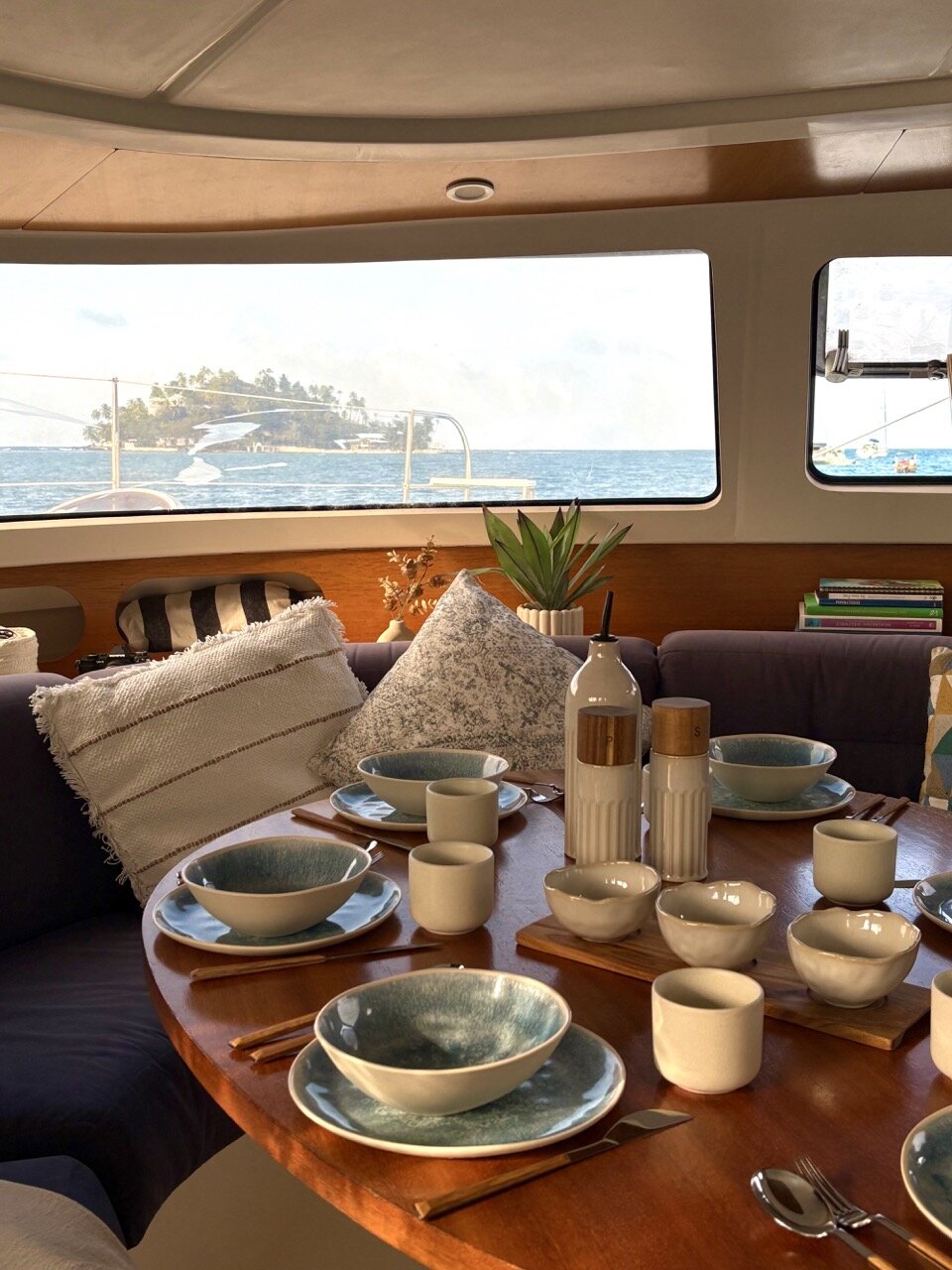Interior view of SV Monika luxury catamaran with a dining table set for a meal, decorative vases, and a large window showing the ocean and distant islands.